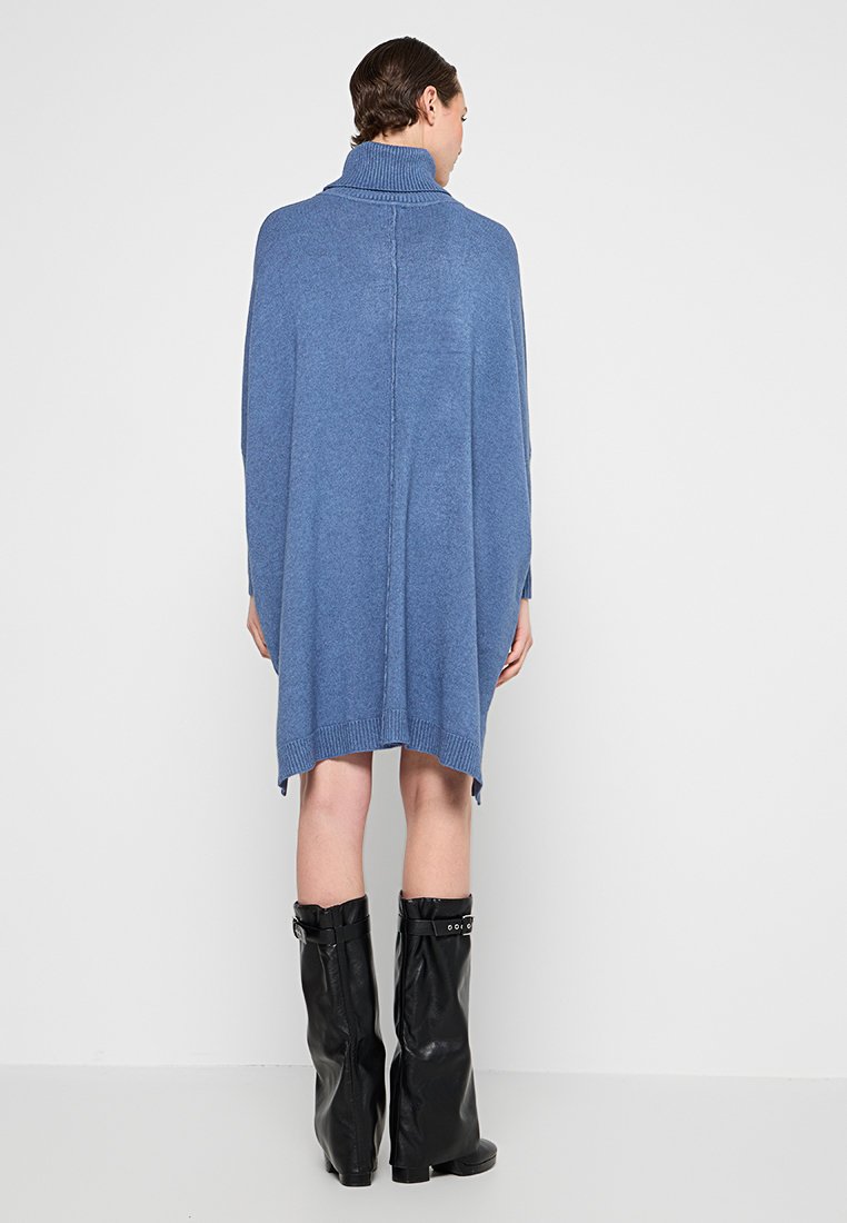 Blau gestrickter Poncho mit hohem Kragen, seitlichen Schlitzen und gerippten Details. Das Model trägt schwarze knielange Stiefel im Kontrast.