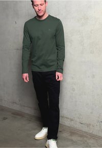 Zielona bluzka z długim rękawem uszyta z miękkiego materiału, w połączeniu z czarnymi spodniami i białymi trampkami z zielonymi akcentami, na tle betonu.