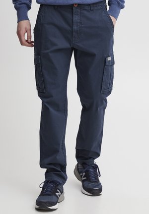 Homme portant un pantalon cargo bleu marine avec poches latérales et des baskets bleues marines, debout devant un fond clair uni.