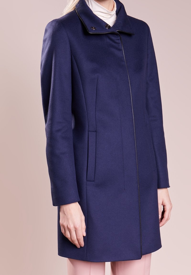 Femme portant un manteau bleu marine longueur genou avec un col montant et des poches latérales, par-dessus un pull à col roulé blanc et un pantalon rose clair.