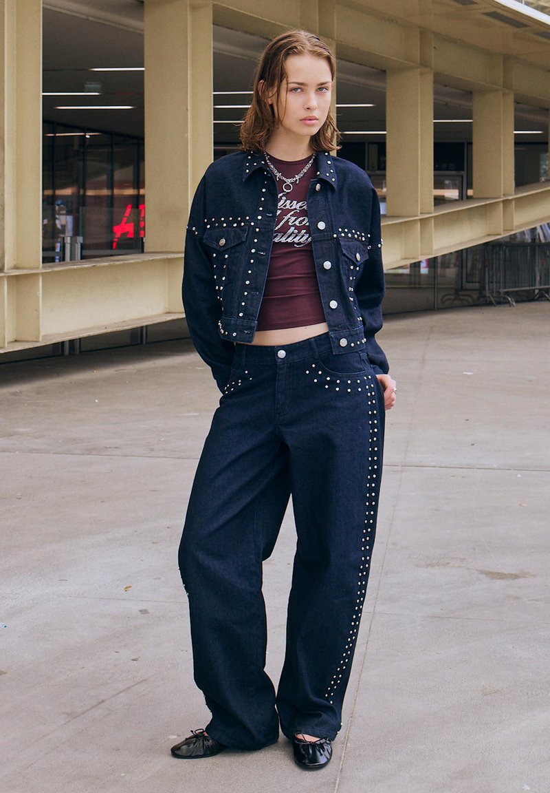 Denim jakna in hlače v temno modri barvi z dodatkom srebrnih žebljičkov vzdolž jakne in hlač. Model nosi burgundski top in črne balerinke.