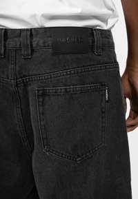 Mustat denim-farkut mattapinnalla, joissa on suuri nahkainen vyötärötunnus, johon on painettu "WASTED", sekä pieni brändätty tunnus sivutaskussa.