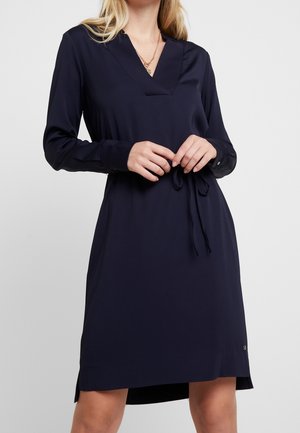 Marineblauwe jurk met een V-hals, lange mouwen en een strikceintuur in de taille. Gladde stof met zijsplitten in de zoom en een klein logodetail.