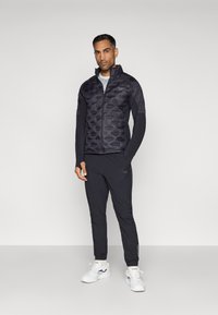 Μαύρο μπουφάν με επένδυση και πικέ σχέδιο, με ψηλό γιακά, συνδυασμένο με μαύρες αθλητικές παντελόνες και λευκά αθλητικά παπούτσια. Διακριτές λογότυπα.