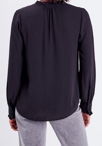 Blusa negra de manga larga con cuello con volantes y puños abotonados. Tejido suave con un corte relajado, que presenta un sutil brillo y sin patrones visibles.