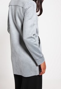 Chaqueta de ante sintético gris claro con cuello de muesca, cierre de un solo botón y bolsillos laterales, que presenta una textura suave y un corte relajado.