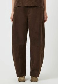 Pantaloni a gamba larga in camoscio marrone con una texture liscia, caratterizzati da dettagli a pannelli cuciti e una vestibilità comoda. Abbinati a stivaletti marrone chiaro.