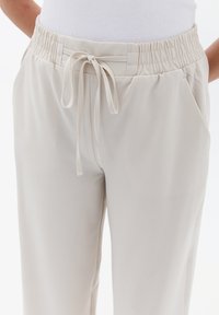 Beige kalhoty s elastickým pasem a šňůrkou, které mají boční kapsy a hladkou, texturovanou látku.