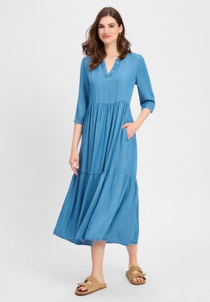 Jeansmaxikleid mit drei Viertel Ärmeln, V-Ausschnitt, mehrlagigem Design und seitlichen Taschen. Ergänzt durch hellbraune flache Sandalen.
