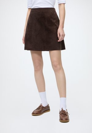 Person iført en mørkebrun ruskindsnederdel, hvide sokker, brune båd shoes og en hvid top, stående mod en ensfarvet lys baggrund.