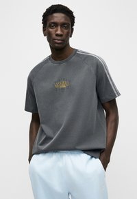 Γκρι βαμβακερό μπλουζάκι με κοντά μανίκια, στρογγυλή λαιμόκοψη και χρυσό γράφημα "Originals". Συνδυασμένο με ανοιχτό μπλε φόρμα για χαλαρή εμφάνιση.