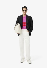 Vita sneakers, vita byxor, en svart kavaj och en rosa randig polo. En beige väska hålls i ena handen; solglasögon fulländar looken.