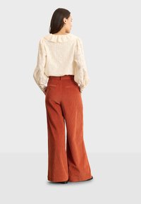 Blusa bianca con texture e colletto arricciato, abbinata a pantaloni in corduroy a gamba larga color ruggine con tasche posteriori e cuciture visibili.