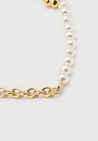 Τμήμα αλυσίδας από χρυσό συνδεδεμένο με μια σειρά στρογγυλών λευκών μαργαριταριών σε λευκό φόντο.