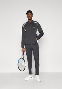 Conjunto atlético masculino em preto e branco. Inclui uma jaqueta com fecho de correr e logotipo, calças ajustadas e ténis brancos. Segura uma raqueta de ténis.