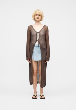 Kvinde med krøllet blondt hår iført en lang brun gennemsigtig strikket cardigan over en hvid top, lys blå asymmetrisk denimnederdel og sorte sandaler.