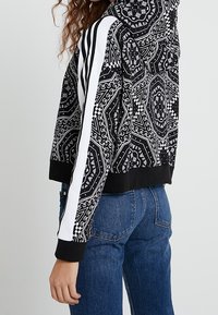 Černá krátká mikina s kapucí a složitými bílými vzory, s kapucí, bílými bočními pruhy a žebrovanými manžetami. Kombinována s modrými džíny s vysokým pasem.