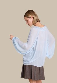 Lyseblå chiffonbluse med flagrende ærmer, samlet halsudskæring og en blød drapering. Sat sammen med en brun plisseret nederdel. Neutral baggrund.