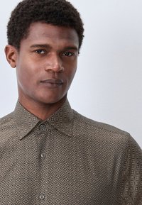 Camicia marrone fantasia con motivi geometrici, colletto con bottoni e tessuto dalla texture liscia; bottoni visibili sulla parte anteriore.