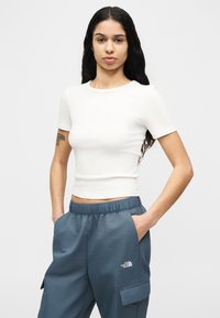 Γυναίκα με μακριά σκούρα μαλλιά, φορώντας εφαρμοστό λευκό κοντομάνικο τοπ και μπλε παντελόνι καμπαρντίνα με το λογότυπο The North Face, με τα χέρια στις τσέπες.