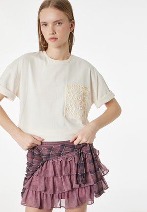 Młoda kobieta z długimi włosami, nosząca duże kolczyki koła, kremową koszulkę z krótkim rękawem z haftowaną kieszenią oraz fioletową spódnicę z warstwowym wzorem.