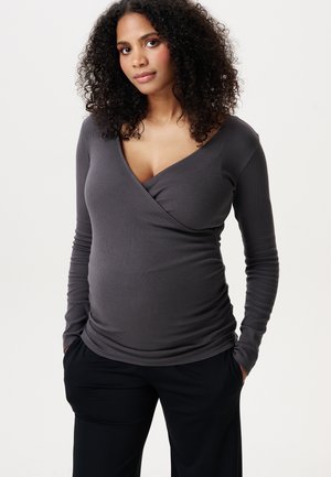 Vrouw met krullend haar die een donkergrijze lange-mouwen overslagtop en zwarte broek draagt, staand met handen in de zakken tegen een effen achtergrond.