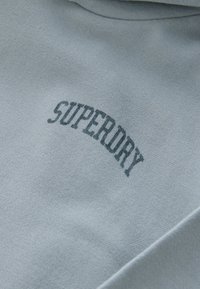 Hele sinine fliistega dressipluus kaarekujulise halli "SUPERDRY" logoga. Pehme tekstuur, õrn läige ja minimalistlik disain ilma lisakaunistusteta.