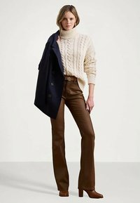 Moteris vilkinti kreminės spalvos mezginio aukštu kaklu megztinį, rudos odos kelnes, rudus batus ir laiko tamsiai mėlyną paltą per vieną petį.