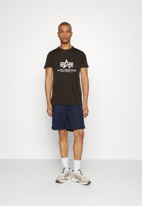 Mørkebrun t-shirt med hvid "Alpha Industries" logo, parret med marineblå shorts, hvide sokker og beige sportssko med sorte detaljer.