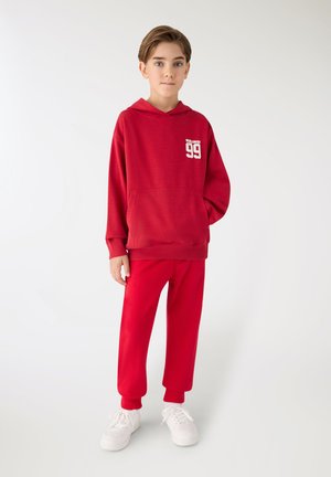 Ragazzo in piedi che indossa una felpa rossa con cappuccio con la scritta "Worldwide 99", pantaloni rossi e sneakers bianche su sfondo chiaro uniforme.