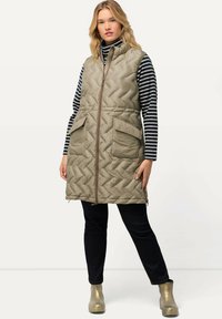 Pikowana, oliwkowo-zielona kamizelka z wysokim ko łnierzem, zamkiem błyskawicznym z przodu i dwiema dużymi kieszeniami. Do stroju dopełniają koszula w paski i ciemne spodnie.