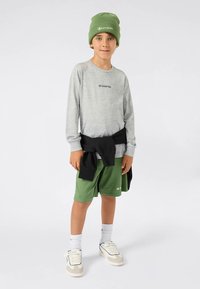 Fiú áll sportos Champion öltözetben, zöld sapkában, szürke hosszúujjú pólóban, zöld rövidnadrágban, fehér tornacipőben, és fekete pulóverrel a derekán.