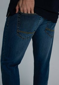 Granatowe dżinsy o gładkiej fakturze, z podwójnym przeszyciem i dwoma tylnymi kieszeniami. Widoczny detal ręki w kieszeni.