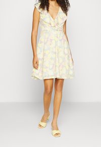 Robe fleurie aux tons pastel jaune, bleu et rose avec un col à volants et des manches courtes. Silhouette trapèze, longueur genou, portée avec des sandales à talons jaunes.