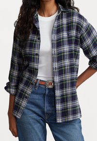 Rutig flanellskjorta i marinblått, grönt och gult, med knäppning fram, långa ärmar, och bärs över en vit t-shirt. Blåjeans och bälte synliga.