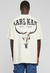 Krämfärgad t-shirt med kort ärm som har en stor grafisk design av en tjurhornskalle och texten "KARL KAN EST. 1989" i sliten svart bokstavsform.