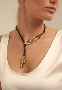 Femme portant un collier en cordon noir avec un grand pendentif incurvé en or et trois charms en forme de goutte d'or sur un haut crème.