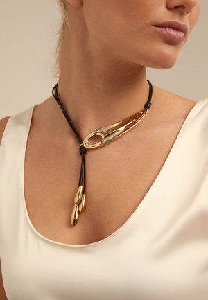 Femme portant un collier en cordon noir avec un grand pendentif incurvé en or et trois charms en forme de goutte d'or sur un haut crème.