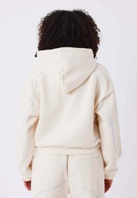Beige Kapuzenpullover mit lockerer Passform, strukturiertem Stoff, kontrastierenden blauen Nähten und verstellbarem Kapuzenzug. Sichtbare Nähte und elastische Bündchen.