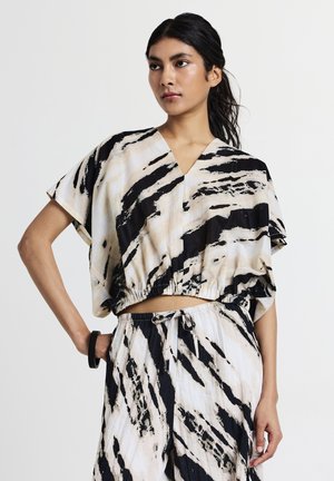 Une jeune femme aux longs cheveux noirs porte un haut ample à rayures noires et blanches et un pantalon à cordon assorti, posant avec une main sur la hanche.