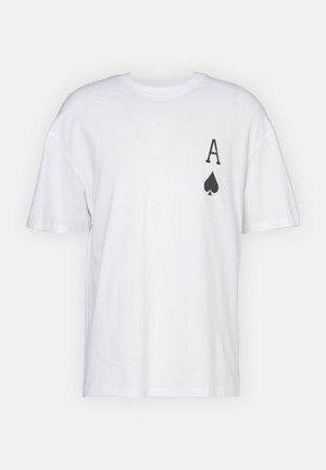Mand iført en hvid T-shirt med et sort "A" og spar-symbol på brystet, parret med løse brune jeans, stående mod en ensfarvet baggrund.