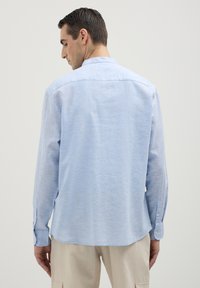 Uomo che indossa una camicia a maniche lunghe azzurro chiaro e pantaloni beige, girato di spalle contro uno sfondo grigio chiaro uniforme.
