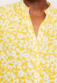 Blouse boutonnée à fleurs jaunes et blanches avec un col à volants portée par une personne à la peau foncée.