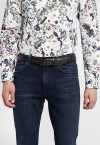 Cintura in pelle marrone con fibbia regolabile, indossata su jeans in denim blu scuro; abbinata a una camicia bianca con motivo floreale.