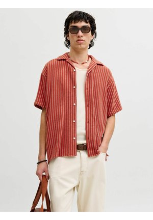 Jeune homme portant des lunettes de soleil, chemise à manches courtes rayée rouge et blanc sur un débardeur blanc, pantalon crème, tenant un sac marron devant un fond uni.