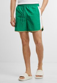 Shorts de bain verts avec des panneaux latéraux blancs, taille élastique et cordon de serrage. Présente des finitions en piping beige le long des ourlets. Le tissu a l'air léger.