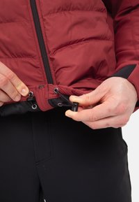 La giacca a sacco rossa presenta una zip e un orlo regolabile con cordino, mostrando una texture trapuntata e un rivestimento nero a contrasto. Mani che tengono l'orlo.
