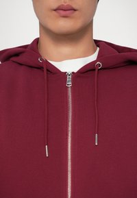 Hoodie bordeaux à fermeture éclair argentée. Doté d'une capuche avec cordon de serrage et d'un col intérieur blanc, fabriqué à partir d'un tissu doux et texturé.