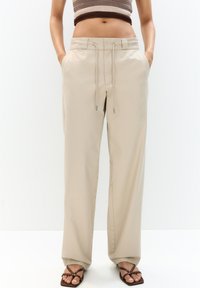 Pantalons larges beiges avec une taille à cordon, des poches latérales et une texture lisse, associés à des sandales brunes.