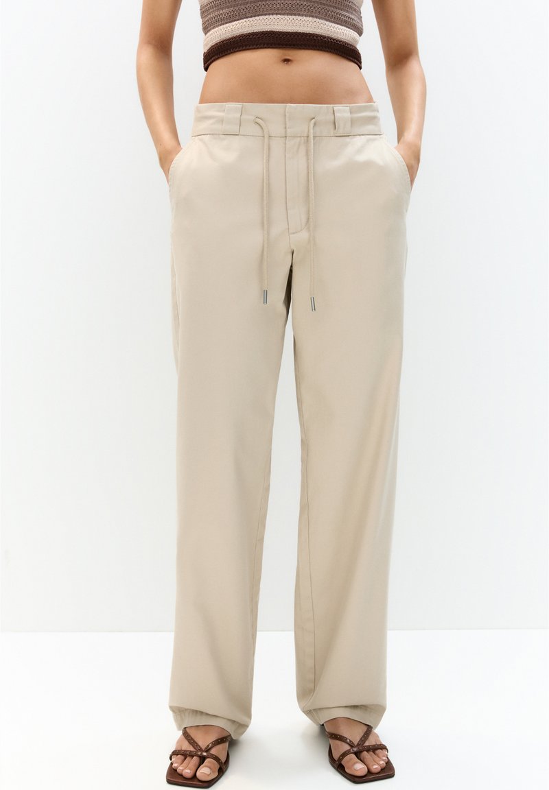 Pantalons larges beiges avec une taille à cordon, des poches latérales et une texture lisse, associés à des sandales brunes.